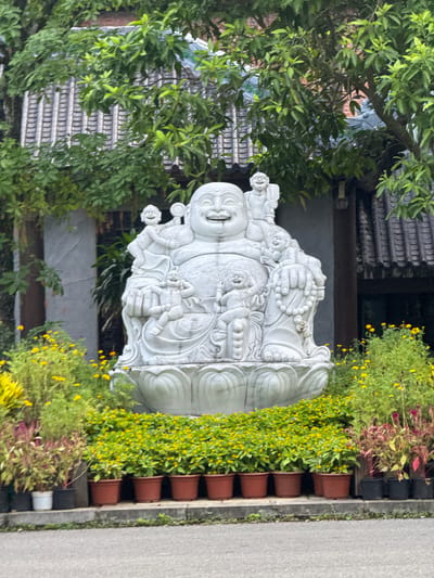 In Tây Hoa Lư Ward, Vietnam, a stone statue of a laughing Buddha, also known as Budai, is prominently displayed outdoors. The statue depicts a plump, jovial figure seated on a lotus pedestal, surrounded by several cherubic children. The Buddha statue is the central element of the scene, positioned in front of a building with a traditional tiled roof and lush green trees. The foreground is adorned with a vibrant arrangement of potted flowers, including rows of small yellow blooms and some taller plants with pinkish-red foliage. The ground in front of the potted plants is a paved surface, likely a walkway or street. The overall setting appears to be an outdoor space, possibly a garden or a courtyard area of a temple or cultural site.