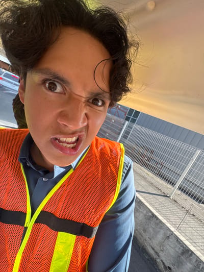 In Sanctorum, Mexico, a male individual is captured in a close-up, front-facing image, exhibiting a pronounced facial expression with wide eyes, a furrowed brow, a scrunched nose, and bared upper teeth. He wears a dark blue collared shirt beneath an orange high-visibility safety vest, which features yellow-green reflective material and black straps. The lighting suggests an outdoor environment with direct sunlight, casting shadows. The upper right portion of the frame shows a light-colored canopy, indicating the subject may be within a vehicle. In the background, a gray building with windows, a white metal security fence, and a concrete ground surface are visible. A white vehicle is partially discernible in the far-left background. The scene depicts an industrial or commercial setting.