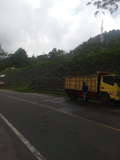 The scene depicts a roadside area in Kandapola, Sri Lanka, under an overcast sky. A two-lane asphalt road, partially wet and featuring a solid white line on the left and a faded double yellow line centrally, occupies the foreground. A yellow and red medium-duty cargo truck is parked on the shoulder to the right of the road, facing left. A male individual, wearing a dark blue shirt and light-colored shorts, stands barefoot beside the truck's passenger side, facing its cargo bed.
Behind the road and truck, a multi-tiered retaining wall, constructed from large dark grey stones or gabion baskets, stretches horizontally across the mid-ground. Green vegetation, including shrubs and trees, grows on and above this wall. Further up the steep hillside, a dense canopy of green trees is visible. A tall, red and white cellular transmission tower is prominent on the upper right side of the hillside, partially obscured by foliage. The sky is largely overcast with grey clouds, suggesting either an impending rain or recent precipitation, consistent with the wet patches on the road surface. A small, multi-colored umbrella is partially visible on the left side of the retaining wall, near the road.