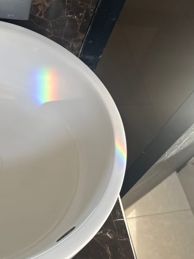 A close-up, high-angle shot captures a white sink with a faint rainbow-like light reflection on its surface. The sink appears to be made of porcelain and is positioned in what seems to be a bathroom or washroom setting. The surrounding environment includes dark, marbled countertops and beige tiled flooring, suggesting a modern interior design. The lighting conditions indicate that natural light is entering the space, creating the spectral refraction on the sink. There are no people or visible activities taking place in the image. The location in Barquisimeto, Venezuela, cannot be determined from the visual information provided in the image.