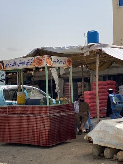 The photograph shows a street scene in Arghandab, Afghanistan. A food stall is positioned on the side of the road, selling "Karachi Special Chicken Biryani," as advertised in Urdu script above the stall. The stall is covered with a worn white tarp and has a burgundy and brown patterned front. A yellow bucket and other small items are visible on the counter.

A man with a beard is sitting on a blue plastic chair next to the stall, wearing a brown outfit and a white cloth draped over his shoulder. In the background, stacks of red sacks, possibly grain or other goods, are visible behind the food stall. A person wearing a blue jacket and a white turban is standing behind the sacks. A light blue car is parked near the food stall.

The environment appears to be dusty, and the lighting suggests a bright, sunny day. The overall scene depicts everyday life in a busy marketplace setting.