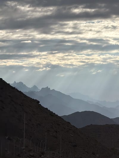 The image depicts a mountainous landscape under a cloudy sky. Sunbeams are visible breaking through the clouds, casting rays onto the distant mountain ranges which fade into a hazy blue silhouette. In the foreground, a dark, rocky slope dominates the lower left of the frame. A fence with posts is visible running horizontally across the bottom portion of this slope. The overall scene conveys a sense of natural grandeur and atmospheric conditions. The provided information about the location in Chumkar, India, is not visually verifiable from the image itself.