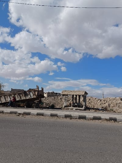 The image depicts an outdoor scene in Al-Qadam Municipality, Syria, characterized by significant urban demolition and debris. In the foreground, a gray asphalt road occupies the lower portion of the frame, bordered by a concrete curb with alternating black and white segments, appearing worn and partially damaged. Beyond the curb, a dirt and debris-strewn shoulder leads into the mid-ground.

To the left in the mid-ground, a large, dark-colored dump truck with its hydraulic bed fully elevated is positioned next to extensive piles of rubble. Two individuals are observed on bicycles: one in a red top and dark pants is riding a dark bicycle near the front of the dump truck, moving from left to right. A second individual, also on a dark bicycle, follows further to the right, behind the dump truck.

To the right of the dump truck, a heavily damaged stone structure with multiple columns, resembling a former classical-style bus stop or shelter, stands amidst the debris. Its roof is partially missing, exposing internal scaffolding or support elements. Behind these foreground and mid-ground elements, vast mounds of earth, concrete rubble, and general debris stretch across the landscape, indicating widespread destruction.

In the background, behind the debris piles, a few damaged building structures are visible, some appearing partially collapsed. On the far right, a slender, light-colored minaret rises above the horizon. The sky is largely clear and blue, interspersed with numerous scattered white cumulus clouds. Two thin, dark utility lines traverse horizontally across the upper part of the sky.