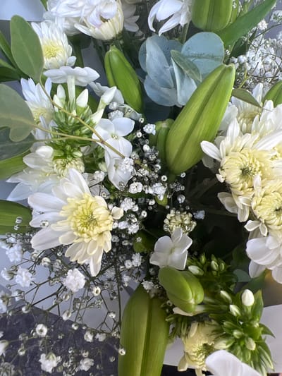 A close-up overhead shot of a bouquet of white flowers and green buds against a dark, speckled surface. The bouquet features large white chrysanthemums with yellow centers, smaller white star-shaped flowers, and delicate white baby's breath. Large, unopened green buds of what appear to be lilies are also prominent. Sprigs of gray-green eucalyptus leaves are interspersed throughout the arrangement. The background surface is dark and textured, possibly a table or countertop. The lighting is soft and diffused, creating a gentle illumination of the floral subjects. There are no people visible in the image. The image does not provide any information about the location of Hadera, Israel.