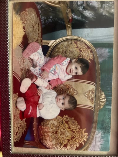 Two infants are depicted lying on a red and gold patterned sofa or chaise lounge within an indoor setting in Ksar-el-Kebir, Morocco. The image is a physical photograph contained within a red, textured album frame, slightly rotated clockwise.

The upper-right infant, appearing slightly older, is positioned on its back, head turned towards the camera with an open-mouthed smile. This infant is dressed in a light pink long-sleeved top featuring a cartoon character on the chest, patterned white and pink trousers, and pink shoes with textured soles.

The lower-left infant is also on its back, head turned towards the camera, maintaining a neutral expression. This infant wears a white knitted long-sleeved sweater, red trousers, and light-colored soft shoes or socks.

The furniture features ornate gold-colored wooden frames and deep red upholstery with intricate gold and brown floral or damask patterns. A blurred background indicates additional furniture or room elements, possibly a wall or curtain with a light teal or green hue, and reflections are visible on the photograph's surface.