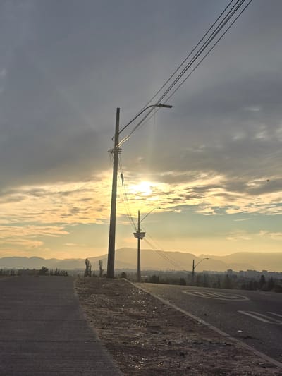The image shows a street in Puente Alto, Chile, at dawn or dusk. In the foreground, there is a gray asphalt road with a dirt shoulder. White markings are visible on the road, including what appears to be a circular speed limit symbol. To the left, utility poles with power lines stretch across the sky. The background contains a cityscape nestled against mountains, partially obscured by a golden sky filled with clouds, through which the sun is shining. There are no people visible.