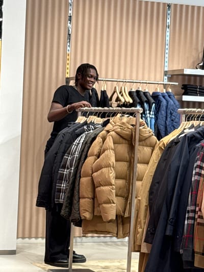 In a retail clothing store in La Roca del Vallès, Spain, a young Black man with dreadlocks is seen browsing a rack of puffer jackets. He is wearing a black t-shirt and black pants. The store features beige, pleated curtains as a backdrop and displays a variety of outerwear including puffer jackets in brown and blue, as well as plaid jackets. The man is looking at the clothes and appears engaged with the selection.