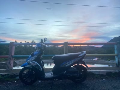 The image depicts a serene twilight scene with a blue and white Yamaha Mio scooter parked on a gravel surface. The scooter is positioned on the right side of the frame, facing left. Beyond the scooter, a low-lying concrete barrier with colorful railings runs horizontally across the mid-ground. A body of water, reflecting the sky, is visible behind the barrier. In the distance, lush green hills and palm trees are silhouetted against a sky filled with pastel hues of pink, orange, and blue, indicative of a sunset or sunrise. The overall atmosphere is tranquil. This scene is located in Lambaro Kueh, Indonesia.