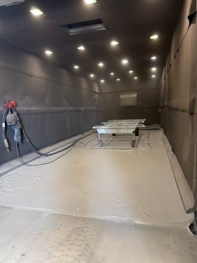 The image shows an interior view of a large, industrial spray booth, likely located in Mol, Belgium. The booth features a dark gray, seamless wall and ceiling construction with numerous square LED lights illuminating the space. The floor is covered in a light gray, dusty material, possibly overspray or sand, with visible tire tracks and footprints. In the center of the booth, a wheeled metal cart holds several rectangular, silver frames. To the left, attached to the wall, is a piece of industrial equipment with hoses and a helmet, suggesting active use for painting or coating. The overall impression is of a functional, well-lit workspace dedicated to industrial finishing processes.