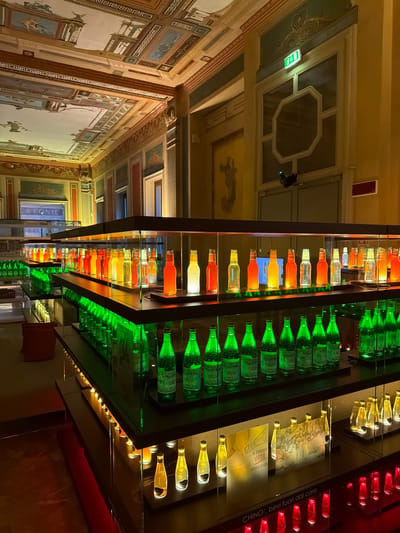 The image depicts an interior view of the Galleria Arte Contemporanea at the San Pellegrino Terme, Italy. The room is elaborately decorated with ornate ceilings featuring painted murals and intricate molding. The primary subjects are rows of glass bottles displayed on illuminated shelves within modern glass cases.

The bottles are varied in color, with prominent displays of green, orange, yellow, and clear glass. The green bottles, some labeled "S. Pellegrino," are arranged in rows on lower shelves, illuminated from beneath with green light, creating a vibrant glow. Above them, on higher shelves, are rows of bottles in shades of orange, yellow, and clear, lit with warm, yellow light, casting a dramatic effect. Some of these bottles appear to contain liquid of varying colors.

In the foreground, a dark shelf displays a row of red illuminated bottles, and above that, a shelf with yellow illuminated bottles. To the right, two informational panels with images are placed on a shelf. One panel features text that reads "la limonata vera sempre pronta."

The environment suggests a museum or exhibition space. The walls are painted in warm tones and feature additional decorative elements, including painted figures and architectural details. An illuminated green exit sign is visible on the upper right side of the image, mounted above a large window or glass door. A black security camera is positioned above this doorway. The overall lighting is dramatic and focused on the displays, with the ornate architecture providing a historical backdrop to the contemporary exhibition. No people are visible in the image.