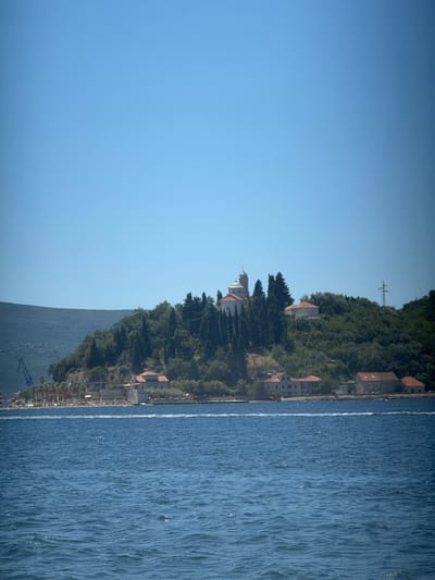 The image captures a serene coastal scene in Lepetane, Montenegro. A clear, bright blue sky dominates the upper portion of the frame. Below, a lush green hillside slopes down towards the water. Nestled on the hillside are several buildings with red-tiled roofs, and a prominent white church with a green dome and a bell tower stands out. A cluster of cypress trees and other greenery surround the structures. The foreground features the deep blue water of the sea, with gentle ripples indicating a calm day. Along the shoreline, more buildings and some palm trees are visible. On the far left of the image, a large construction crane is positioned, suggesting ongoing development or infrastructure work in the area. The overall impression is one of a tranquil, sunny day in a picturesque coastal town.