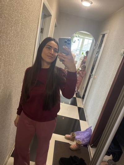 This image depicts an indoor hallway or entryway in the city of Устиново, Russia, captured as a mirror selfie. The primary subject is an adult female with long, dark brown hair, wearing a dark maroon long-sleeved sweatshirt featuring a dark emblem, and light pink trousers. She holds a silver-colored smartphone with a multi-lens camera system in her right hand, with a white nail polish visible. A ring is present on her left ring finger.
Reflected in the mirror, further down the hallway through an arched doorway, a young child with light hair is visible, dressed in a light pink two-piece outfit. The child appears to be looking downwards. Partially visible behind the child is another adult figure, in a reddish garment, facing away from the mirror.
The environment includes walls covered in a textured light beige wallpaper. The floor in the foreground consists of black and white checkered tiles, transitioning to a dark doormat. On the floor near the mirror, a pair of light pink, low-cut, insulated boots and a pair of dark-colored fuzzy slippers are present, alongside a crumpled translucent purple plastic bag. A round, flush-mount light fixture is on the ceiling. Two doors, one white and one dark brown, are visible in the reflection. Beyond the arched doorway, a brightly lit area with what appears to be a window and some green foliage is discernible.