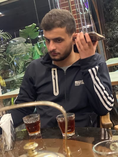 A young adult male, estimated to be in his late teens or early twenties, with short, wavy dark hair and a short beard, is seated at a table. He is wearing a dark blue Adidas track jacket with white stripes on the left sleeve and a white-outlined zippered chest pocket, over a grey t-shirt. He holds a gold-colored smartphone to his left ear with his left hand, appearing to be engaged in a call or listening to audio. His gaze is directed downwards and slightly to his right.
On the dark, reflective table in front of him are two small clear glasses, each containing a dark reddish-brown liquid. A white napkin dispenser is positioned to the left side of the table. A curved metallic object, possibly part of a serving stand, is partially visible in the immediate foreground. The background features dense green foliage, including distinct Monstera leaves, indicating potted plants within an indoor or semi-outdoor setting. Vertical dark elements and subtle reflections of light sources are also present in the background. The lighting appears dim. The specific location within the city of Homs, Syria, cannot be determined from the visual evidence in the image.