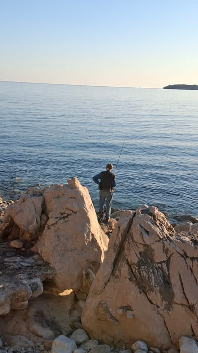 The image depicts a man fishing off a rocky coastline in Podličak, Montenegro. He stands on large, light-colored rocks with a fishing rod extended over the blue water. The sky is clear and light blue, fading to a pale color near the horizon, where a distant landmass is visible. The image is taken at an angle, with the horizon running vertically.