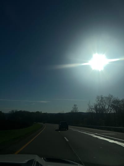 A roadside view, likely taken from inside a vehicle, shows a portion of a road with a yellow line marker. The road is bordered by trees and vegetation. The sky is visible with a bright sun, suggesting daytime. The location is Town of Fishkill, United States of America.