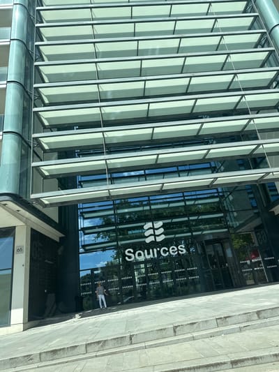 Here is a description of the image.
The image shows the front of a modern building with a glass facade. The building has a sign that reads "Sources" in white lettering, accompanied by a stylized logo. The glass windows reflect the sky and trees. A woman is standing near the entrance. The number "65" is visible on the building's exterior. The building has a series of vertical structures that shade the exterior.
Based on the logo and address number, this is the front entrance of Sources, a company located in Boulogne-Billancourt, France.