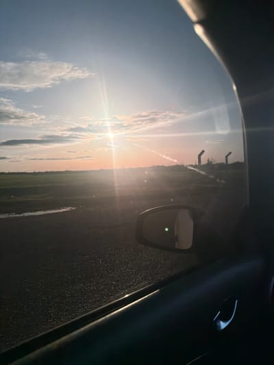 The image depicts an exterior landscape viewed from the interior of a vehicle, specifically through the passenger-side window, in Ait Boubidmane, Morocco. The scene is illuminated by a low sun, positioned near the horizontal center, which emits strong light rays and lens flare, suggesting either sunrise or sunset. The sky transitions from light blue overhead to shades of pale orange and pink closer to the horizon, with scattered cumulus clouds present.

In the immediate foreground, the dark interior trim of a car door panel is visible along the bottom and right edges, including a chrome or silver door handle on the lower right. A side mirror is prominent, showing a reflection that includes a distinct green light point.

Outside the vehicle, a dark, granular road surface extends across the lower left quadrant. Adjacent to the road, a shallow, elongated body of water, possibly a puddle or small stream, reflects the ambient light. Beyond this, a wide, flat expanse of green fields dominates the midground. Along the distant horizon on the right, two dark, L-shaped structural elements, resembling pylons or industrial fixtures, are silhouetted against the sky. No individuals or active subjects are visible within the frame.