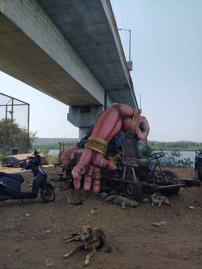 Under a concrete bridge in Chopdem, India, a partially destroyed, large, pink-colored Hanuman statue lies amidst a collection of debris. The statue is missing its head and has elaborate gold-colored armbands. It rests near a dilapidated cart-like vehicle with a steering wheel and seat. Several stray dogs are scattered on the dirt ground nearby, some lying down and others appearing alert. A black scooter is parked on the right side of the image. The background shows a sliver of a body of water with vegetation on the far shore. A partly destroyed fence is in the foreground.