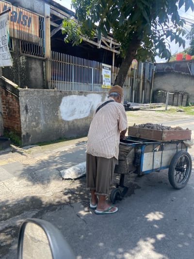 An older man with a light brown striped shirt, brown pants, and sandals stands next to a small, open-air cart containing a pile of dark-colored, irregularly shaped objects. The cart is situated on a paved road in front of a brick building with a metal fence. There is a tree growing on the right side of the image, and the scene is partially shaded by the tree. There is an orange sign with text that appears to be written in Indonesian on the building. The location is in Kecamatan Dayeuhkolot, Indonesia, at latitude -6.963 and longitude 107.594. The man is looking down at the cart, and his right hand is resting on the side of the cart. The scene is taken from a slightly low angle and appears to have been taken with a mobile phone. The man is the only person present in the image.
