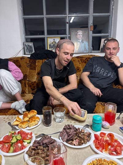 Two men are sitting on a brown patterned couch in a room in Zetiovo, Bulgaria, around a table laden with food. One man wears a black shirt with "UNREAL" printed on it, and he is looking at the camera while his hand rests on his face. The other man, with a partially shaved head and tan scalp cover, is actively using a grater. A third person's feet are visible in the lower-left corner, wearing grey pants and cartoon-patterned socks. The table is covered with plates of tomatoes, meat, fried potatoes, pastries, and beverages in glasses. The room has a window with multiple panes and several framed photographs on the windowsill. The overall lighting in the room is dim.