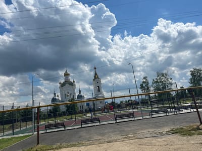 The image depicts an outdoor scene in Dondukovskaya, Russia, under a partly cloudy sky with dramatic, cumulonimbus clouds dominating the upper portion. The time of day appears to be afternoon, given the bright light and shadows.
In the background, a large white church with gold domes and a tall bell tower is prominently visible, suggesting a religious or historical site. In the middle ground, there is a fenced sports court, possibly for basketball or volleyball, with red and green surfacing. Several black benches are placed along the perimeter of the court. A children's playground with red and orange structures is also visible within the fenced area. Tall trees line the back of the sports complex, with more trees and foliage to the right. Power lines stretch across the sky, intersecting with the clouds.
The foreground shows a gravelly path and a section of asphalt, with a yellow railing and a reddish-brown pole in the immediate foreground, part of a fence or sports equipment. The overall impression is of a public space, likely a park or recreational area adjacent to a significant architectural landmark, set against a dynamic sky. There are no people visible in the frame. The weather is bright, but the large clouds suggest potential for rain. No visible text is present in the image.