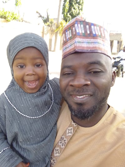 A close-up portrait, likely a selfie, depicts an adult male and a young child outdoors in Babban Giji, Nigeria. The adult male, positioned on the right, has dark skin, a short dark beard with graying hairs, and a slight smile. He wears a light brown traditional top with an embroidered patterned strip over the right shoulder and a multi-colored patterned cap (kufi). The child, positioned on the male's left, appears to be a toddler with dark skin, smiling widely with an open mouth. The child wears a dark gray textured garment with a matching head covering adorned with small white embellishments, and a white beaded necklace. The male is holding the child. The background is brightly lit and blurred, showing light-colored buildings or walls, green foliage, a vertical pole, and a distant figure on a motorcycle to the far right.