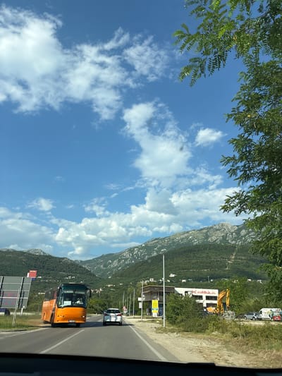 The image depicts a roadside view on a sunny day in Dub, Montenegro, with mountains in the background and a clear blue sky dotted with white clouds. In the foreground, an orange bus is driving on a paved road, followed by a white car. To the right of the road, a yellow excavator is visible next to a building with "PC MRŠULJA" written on it. Trees and vegetation are abundant throughout the scene, with a large tree on the right side of the frame partially obscuring the view of the sky. A speed limit sign is visible on the left side of the road. The overall impression is of a typical summer day in a scenic, mountainous region.