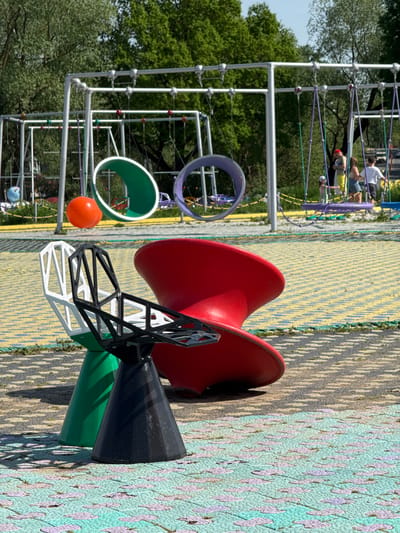 This image shows a playground in the city of Синьково, Russia. The playground features a complex climbing structure with interconnected platforms and rope bridges in the background, where a few people are visible. In the foreground, there's a colorful geometric play structure with red, black, green, and white components. An orange ball hangs on the edge of the climbing structure. The ground is covered with patterned safety mats. The area is surrounded by trees.