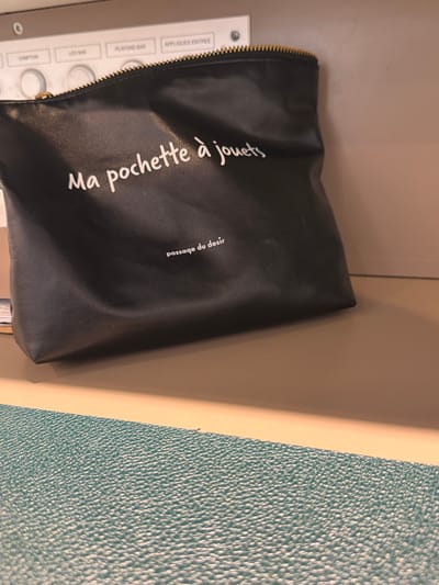 The image shows a black zippered pouch with white text printed on it. The pouch is lying on a surface next to a textured, teal-colored object. Behind the pouch and the teal object, there is a light brown surface.

The text on the pouch reads "Ma pochette à jouets" on one side and "passage du desir" on another smaller section of text. Below the pouch, part of a white label with some text is visible, suggesting it might be attached to a piece of furniture or equipment. The label has some button-like indentations and what appear to be circular cutouts.

The lighting suggests an indoor setting, possibly a room or a closet. The image is shot from a slightly elevated angle, looking down at the objects. The overall impression is one of a personal item placed in a storage area. The time of day and weather are not discernible from the image. The location context is Vienne, France.