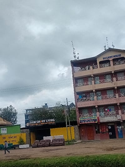An urban street scene in Nanyuki, Kenya, captured on an overcast day, displays commercial establishments and a residential building. In the mid-ground right, a multi-story building, identified by a rooftop sign as "SG LIGHT HOUSE," features beige and reddish-brown facades with decorative balcony railings. Clothes are visible drying on some balconies. The rooftop is equipped with multiple antennae. At street level on this building, storefronts include "PABLO'S BARBER STORE" and "NOONAK ENTERPRISES." Two ground-level doors, one reddish-brown and one black, are present, alongside a partial view of blue vending or ATM machines. To the left of the "SG LIGHT HOUSE" building, an outdoor furniture display features several brown and white/light-colored sofas and couches arranged on an unpaved surface. Behind this display, a darker building is partially obscured by a yellow barrier, bearing a large sign that reads "DIEM SMART LIVING FURNITURE & HOME APPLIANCES." Further to the left, a green structure with a sign for "IDEAL FURNITURE HOME AND OFFICE" is visible. In the foreground, a single adult male, wearing a dark shirt and light trousers, walks from left to right on an unpaved path. Utility lines traverse the upper portion of the scene. The immediate foreground consists of a strip of green vegetation.