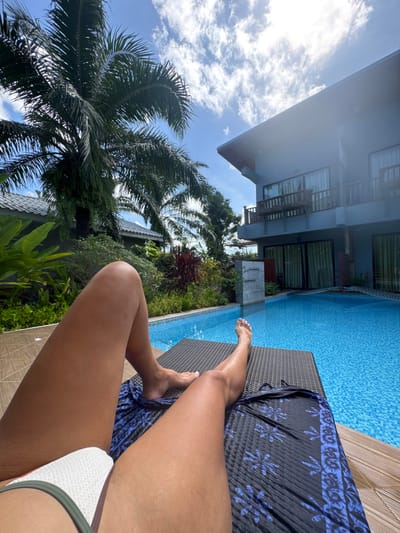 A person is lying on a pool chair, with a blue flowered towel. The person's legs are visible, and they appear to be wearing a bathing suit. Beside the pool chair is a bright blue pool, lush green plants, and a palm tree. The sky is light blue with some white clouds. A building with balconies is in the background. The photo is taken at an unusual angle.