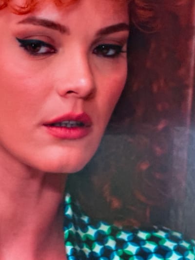 The image is a close-up portrait of a woman, focusing on the left side of her face. She has auburn hair styled in loose curls, and her skin is illuminated by a warm, pinkish light. Her left eye is visible, adorned with dark eyeliner and mascara, and her eyebrow is neatly shaped. The woman is wearing a patterned garment, predominantly green and blue, which is partially visible in the lower right portion of the image. The background is dark and out of focus, with hints of reddish-brown tones, suggesting an indoor setting. The provided location of Uyutnoye, Ukraine, is not discernible from the visual information in the image itself.