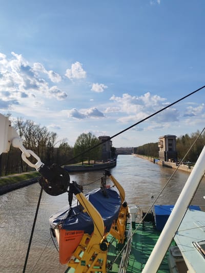 The image shows a view from a boat navigating a canal in Водник Снт, Russia. The canal is flanked by stone buildings with trees lining the banks. In the foreground, parts of the boat are visible, including a yellow lifeboat hoist and an orange lifeboat. The water in the canal is murky brown. A metal support extends from the boat and a black cable is strung across the canal. The sky is blue with scattered white clouds.