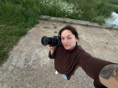 A medium shot shows a person standing on a gravel path near a body of water and lush green vegetation. The person, holding a black Canon EOS 6D camera with a lens attached, is looking directly at the camera. They are wearing a brown long-sleeved shirt, blue jeans, and white sneakers. A tattoo is visible on the person's left arm. The background features a concrete embankment, tall grass, and white wildflowers. The setting appears to be an outdoor environment, possibly a park or natural area within Большая Уса, Russia.