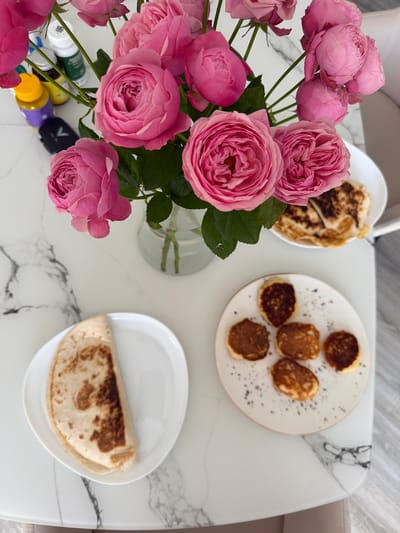 On a white marble-patterned table, a vase of pink roses sits next to a plate of mini pancakes and another plate with a folded tortilla. Several small bottles and a black electronic device are visible behind the roses. The scene appears to be set indoors, possibly in a home in Malinovka, Russia, though this location cannot be definitively confirmed from the image.
