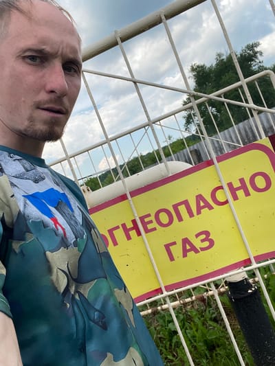 A close-up, low-angle selfie captures a man with light skin, sparse facial hair, and a neutral expression, looking towards the camera. He is positioned in front of a white metal fence. His dark green t-shirt features a camouflage pattern with an emblem on the left chest depicting the Russian flag. Behind the fence, a yellow sign with a pink border is visible, bearing the Cyrillic text "НЕОПАСНО" and "ГАЗ", which translates to "Dangerous" and "Gas". Beyond the sign, a white, cylindrical object, likely a gas tank, and lush green vegetation are present under a cloudy sky. The setting appears to be an outdoor industrial or utility area in Nizhnii Bugrysh, Russia.