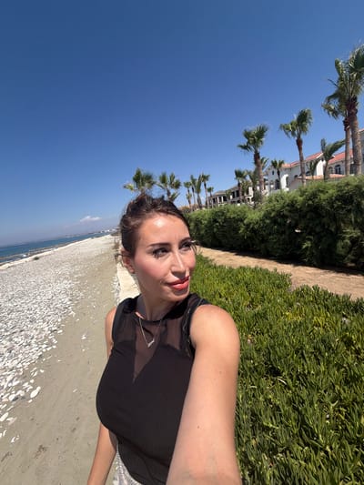 A woman with her hair pulled back is taking a selfie on a sunny day in Pervolia, Cyprus. She is wearing a black top with a sheer neckline and a silver necklace. She is standing on a path next to a beach with white pebbles and the sea on her left. Behind her, there is a line of lush green bushes and palm trees, with white buildings in the background. The sky is clear and blue.