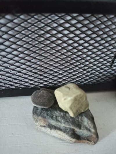 A close-up, eye-level shot shows three stones balanced on top of each other against a light-colored, textured surface. Behind the stones, a black metal mesh with diamond-shaped openings is visible, suggesting it is part of a grate or screen. The stones are positioned in the foreground, with the largest, a dark grey and tan striped rock, at the bottom. On top of this, a small, round, dark grey stone is balanced, and a pale, irregular-shaped stone rests on top of that. The lighting is diffused, creating soft shadows. The image does not provide enough context to determine its location in Promyshlennaya, Russia.
