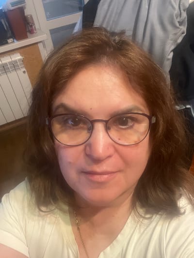 A close-up, selfie-style portrait of a woman with medium-length brown hair and glasses is captured indoors. She is wearing a light-colored t-shirt and a gold necklace. The background shows a radiator and a window, suggesting a residential setting. The lighting appears to be artificial, likely from an indoor source. The woman is looking directly at the camera, with a neutral expression. There are no visible signs of the city of Tula, Russia, in the image.