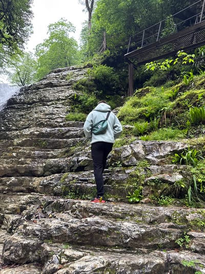 The image shows a person standing on a layered rock formation in a natural environment. The person is wearing a light-colored hooded sweatshirt, black pants, and athletic shoes with red accents, with a green bag slung across their back. They are positioned facing away from the camera. The rock formation is characterized by vertical striations and is partially covered with moss and various green plants. To the left of the rock formation, there is a view of trees with green foliage, suggesting a forest or wooded area. Above the person and to the left, there is a metal structure, resembling a bridge or platform, partially obscured by trees. The bottom left corner suggests the presence of a waterfall, based on the blurred water. The location is identified as Tsabal, Abkhazia, though specific landmarks are not visible.
