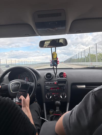 The image provides a driver's perspective from inside an Audi car, looking out onto a highway under a partly cloudy sky during daytime. The scene suggests the car is in motion.
In the foreground, a person's right hand, presumably the driver's, is holding a smartphone that rests on the lower section of the Audi-branded steering wheel. The phone's screen displays a music player interface with track titles such as "Moke - El Paraiso e a dor..." and options for radio genres like "Pop". To the far right, the shoulder and upper torso of another individual, wearing a dark grey t-shirt, are visible. The rearview mirror reflects a partial view of a person.
The car's dashboard is equipped with an Audi Concert radio and climate control system. Visible text on the display includes "FM1 108.0", "28.0 LO" for fan settings, and an external temperature of "17.0°C". Small decorative items hang from the rearview mirror, including a yellow figure with "SMILES" text and a miniature motorcycle. Two red figurine decorations are positioned on the dashboard. A white charging cable is seen connected and resting near the passenger's seat. Keys are visible near the gear shift.
Outside the vehicle, a multi-lane road is discernable, bordered by a tall sound barrier or fence on the left, indicative of a highway setting. The sky above is bright with scattered clouds. This journey takes place in a location consistent with Aldomirovtsi, Bulgaria. The act of holding and interacting with a smartphone while operating the vehicle is a prominent detail.