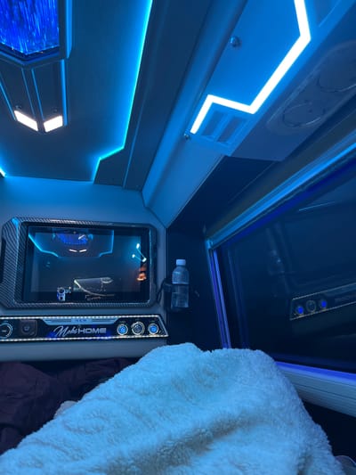 The image shows the interior of a bus or luxury vehicle, likely in Dong Nai City, Vietnam, illuminated by vibrant blue LED lighting. The scene is detailed and features several key elements.
The ceiling is adorned with recessed blue LED strips, creating a modern and futuristic ambiance. Above, a rectangular light fixture with vertical blue light elements is visible. There are also air vents integrated into the ceiling.
In the center of the image, a large touchscreen display is mounted. The screen shows an abstract futuristic image with blue accents and appears to be displaying a video or graphic. Below the screen is a control panel with various buttons and labels. Notably, the text "NEW MobiHOME" is prominently displayed, suggesting this might be a mobile entertainment or living space. There are circular buttons, some illuminated with blue light, labeled "SLEEP," "LCD," and "LIGHT." A USB port is also visible on the left side of the control panel.
To the right of the screen, a clear plastic water bottle is secured in a holder. The overall interior suggests a comfortable and technologically advanced environment.
The lower part of the image shows a soft, plush white blanket draped over what appears to be a bed or seating area. Beneath the blanket, a darker-colored quilt or blanket is partially visible, indicating a sleeping or resting arrangement. The background on the right side features a darkened window, reflecting some of the interior lighting. The presence of the "MobiHOME" branding and the luxurious interior suggest this vehicle is designed for travel and potentially overnight stays.