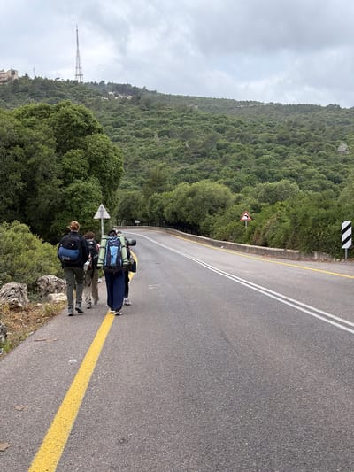 Three people are standing on the side of a paved road in Nesher, Israel. They are wearing backpacks and appear to be hiking or trekking. The road has double white lines and a yellow line along the edge. A mountain covered in green trees is in the background. A tall telecommunications tower is visible on the mountain in the distance. Road signs are present, one indicating a curve in the road. The sky is overcast.