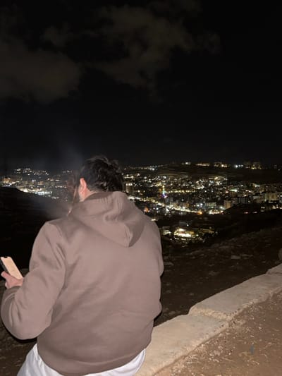 A person stands on the edge of a high cliff overlooking a city at night. The city lights illuminate the landscape. The individual, wearing a brown hooded sweatshirt and white pants, leans forward, peering down into the illuminated cityscape. Clouds are visible in the sky, partially obscuring the dark night. A concrete barrier is to the side of the person, marking the edge of the cliff.
