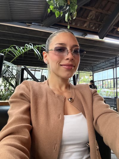 A woman is taking a selfie in what appears to be an indoor dining space in La Tijera, Mexico. She is wearing a beige cardigan over a white top and accessorized with sunglasses and stud earrings. In the background, there are plants and metal framework. The lighting appears to be coming through a glass ceiling or large windows.