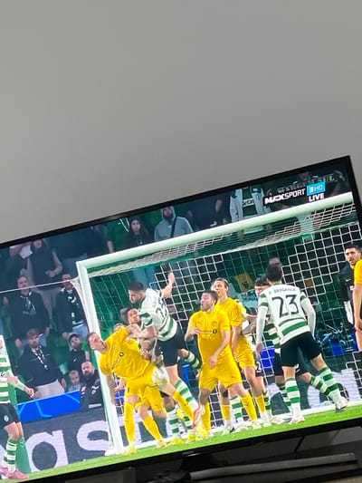This image, captured in Sredkovets, Bulgaria, displays a television screen showing a live football match. The screen occupies the lower two-thirds of the frame, with a plain, light grey wall or ceiling visible above it.

On the television screen, a dynamic moment of a football game is visible around a goal. Players from two teams are in close proximity to a white goal frame with a green net. One team wears white and green vertical striped jerseys with black shorts and striped socks, while the other wears solid yellow jerseys with yellow shorts and socks. A player in a green and white striped jersey, number 30, is airborne near the left goalpost, with his right arm raised and left arm bent. Several yellow-jerseyed players are clustered around him and the goal area, reacting to the play. Another player in a green and white jersey, number 23, identified as "D. BRAGANCA," stands to the right of the goal, observing the action. In the background of the televised match, blurred figures of spectators are visible. On-screen graphics in the upper right corner of the television display "MAXSPORT 3 HD LIVE" and a "BETANO" logo. The television itself appears to rest on a dark, unidentifiable surface.