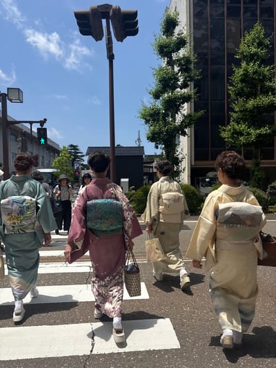 Four women in traditional kimonos are depicted walking away from the viewer, crossing a zebra-marked asphalt street from left to right, in the city of Kanazawa, Japan. The scene is illuminated by bright daylight under a clear blue sky with sparse white clouds.
From left to right, the women are dressed as follows:
1. A woman in a pale teal kimono with a multicolored obi featuring large leaf and floral patterns. She wears white tabi socks and white platform sandals.
2. A woman in a dark pink kimono with a floral print near the hem and sleeves, paired with a shimmering teal and green obi. She wears white tabi socks and light-colored platform sandals.
3. A woman in a light beige kimono with small blue geometric motifs, and a cream-colored obi bearing a red-stamped design. She wears white tabi socks and light-colored sandals.
4. A woman in a pale yellow kimono with an obi depicting a landscape scene including a thatched-roof house and mountains. She wears white tabi socks and brown/gold sandals.
Each of the four kimono-clad women carries a bag: the leftmost carries a light woven bag, the second a dark woven bag, the third a translucent patterned bag, and the rightmost a dark woven basket-like bag.
In the mid-ground, a fifth woman wearing a hat, a patterned top, and dark trousers is visible walking towards the camera on the opposite side of the street.
The street features a white zebra crossing, with a visible crack in the asphalt in the foreground. On the left, a traffic light pole displays a green pedestrian "walk" signal. A larger, dark-colored traffic light pole with multiple light heads stands centrally in the upper frame. Buildings with windows and green foliage from trees line both sides of the street. Shadows of the people and the central traffic light pole are distinctly cast on the street surface.