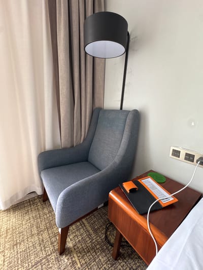 A floor lamp with a black shade stands behind a blue upholstered armchair. The armchair is situated next to a small wooden side table on which a white charging cable is plugged into a wall outlet. On the table, there is a black folder with an orange trim and a green circular object with text. The scene is within a room in Baiyun District, China, indicated by the presence of hotel amenities such as the charging cable and what appears to be a hotel information card or menu on the table, along with a light-colored curtain in the background. The floor is carpeted with a textured brown and tan pattern. The wall is light-colored and shows some minor imperfections.