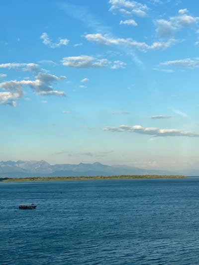 The image depicts a body of water, possibly a lake or sea, with a distant shoreline and mountains visible in the background. The sky above is blue, with scattered clouds. A small boat containing several people is visible on the water. The location is likely near Braćeni, Montenegro, based on the presence of mountains and a large body of water.