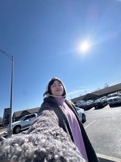 The image captures a selfie-style shot of a person outdoors on a bright, sunny day. The camera is held low, looking upwards, resulting in a composition dominated by a clear blue sky.
In the lower-right foreground, a young person is visible, looking towards the camera with a faint smile. They are wearing a light-colored, possibly off-white or beige, hood or bonnet that frames their face. Beneath the hood, a lavender or light purple knitted sweater is visible. They are also wearing a thick, shaggy grey or dark grey coat that takes up a significant portion of the lower-right side of the frame.
The upper-left and central parts of the image are filled with a brilliant, clear blue sky. A very bright sun with prominent rays is positioned in the upper-left quadrant, creating a strong lens flare effect and indicating intense sunlight.
In the mid-ground, extending from the right side towards the center, there is a paved area that appears to be a parking lot. Several cars are parked in rows, mostly light-colored (white, silver) and dark-colored (black, dark grey) vehicles, including what looks like SUVs or trucks. Behind the cars, a building with a dark roof and dark siding is visible. Further in the distance, some bare trees or shrubs can be seen, suggesting the season might be late fall or winter.
In the lower-left corner, a grey metal pole, possibly a street light or utility pole, rises into the sky. It has an arm-like structure extending from it.
No discernible text is clearly readable on any signs or objects in the image. The overall impression is one of a casual, outdoor snapshot taken during daylight hours with pleasant, clear weather.